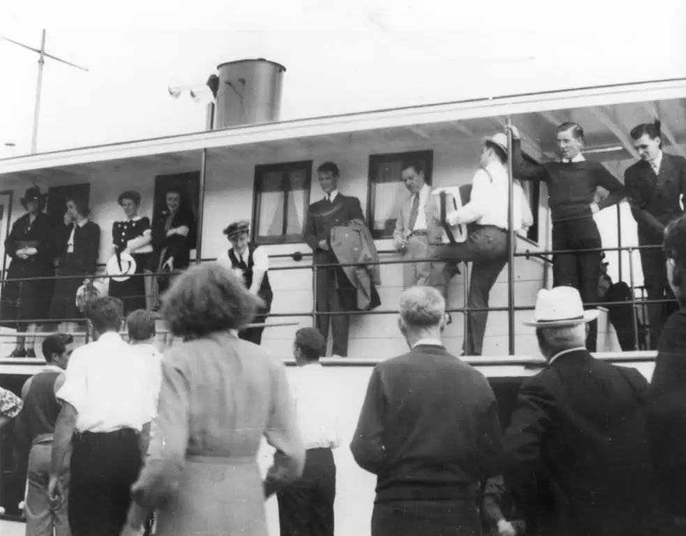 A crowd gathers beside a passenger vessel’s upper deck, where people stand along the railings.