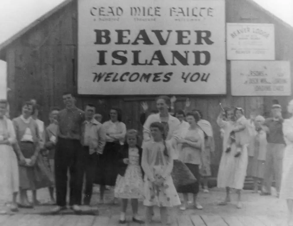 A group of adults and children pose beneath a large welcome sign outside a wooden building.