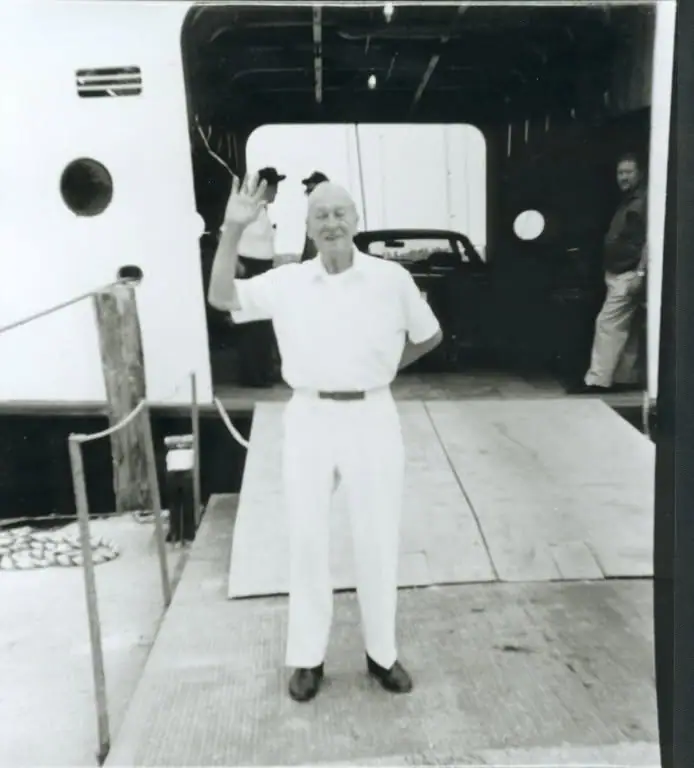 An older man in white clothing waves from a ferry’s vehicle deck.