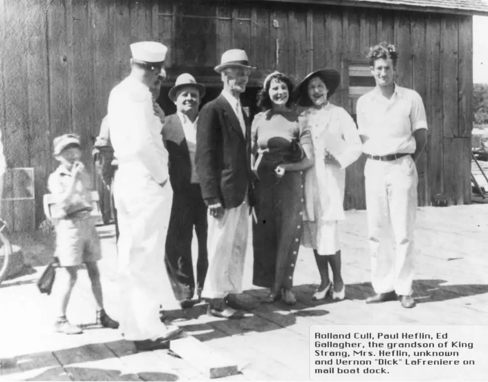 Seven people pose on a wooden dock beside a large wooden building.