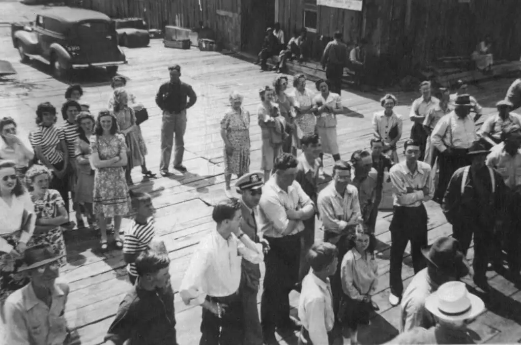 A large group gathers on a wooden pier beside vintage cars and wooden buildings.