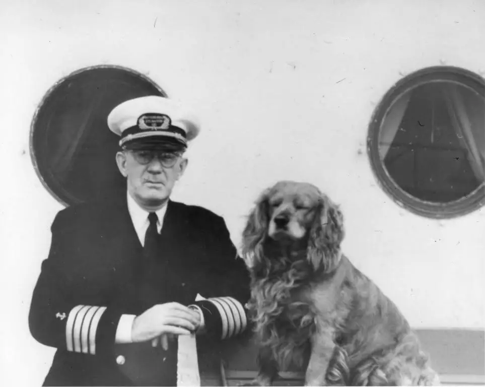 A uniformed man poses beside a long-haired dog against a vessel hull with two round portholes.