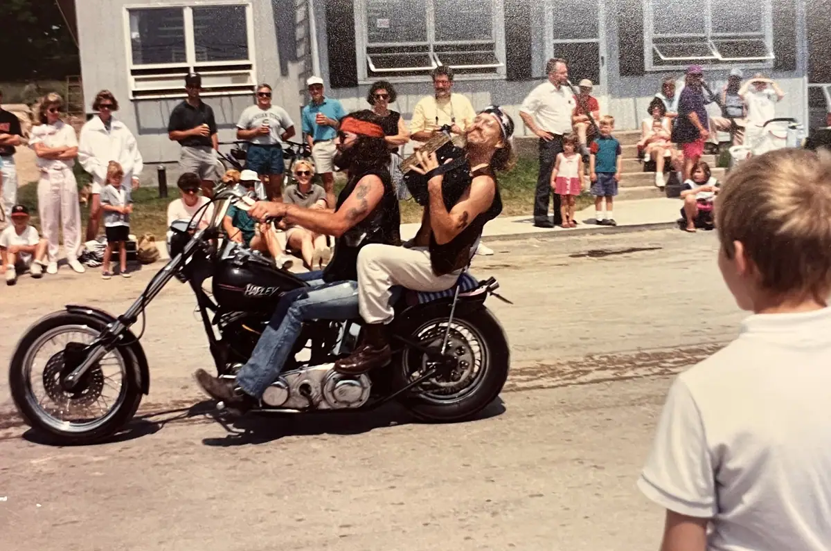 Two adults ride a motorcycle past a crowd gathered outside a building during a public performance.