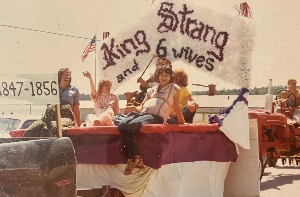A decorated parade float carries several people beside a red tractor, with banners, flags, and historical dates.