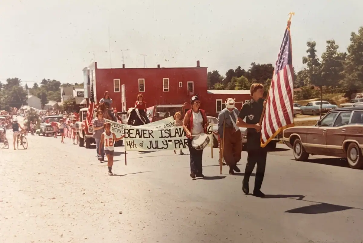 A street parade advances behind a banner, with a flag bearer, drummer, costumed marcher, children, bicycles, and spectators.