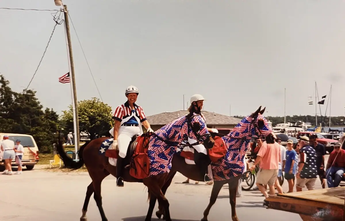 Two helmeted riders lead flag-draped horses past a crowd near boats and a flagpole.
