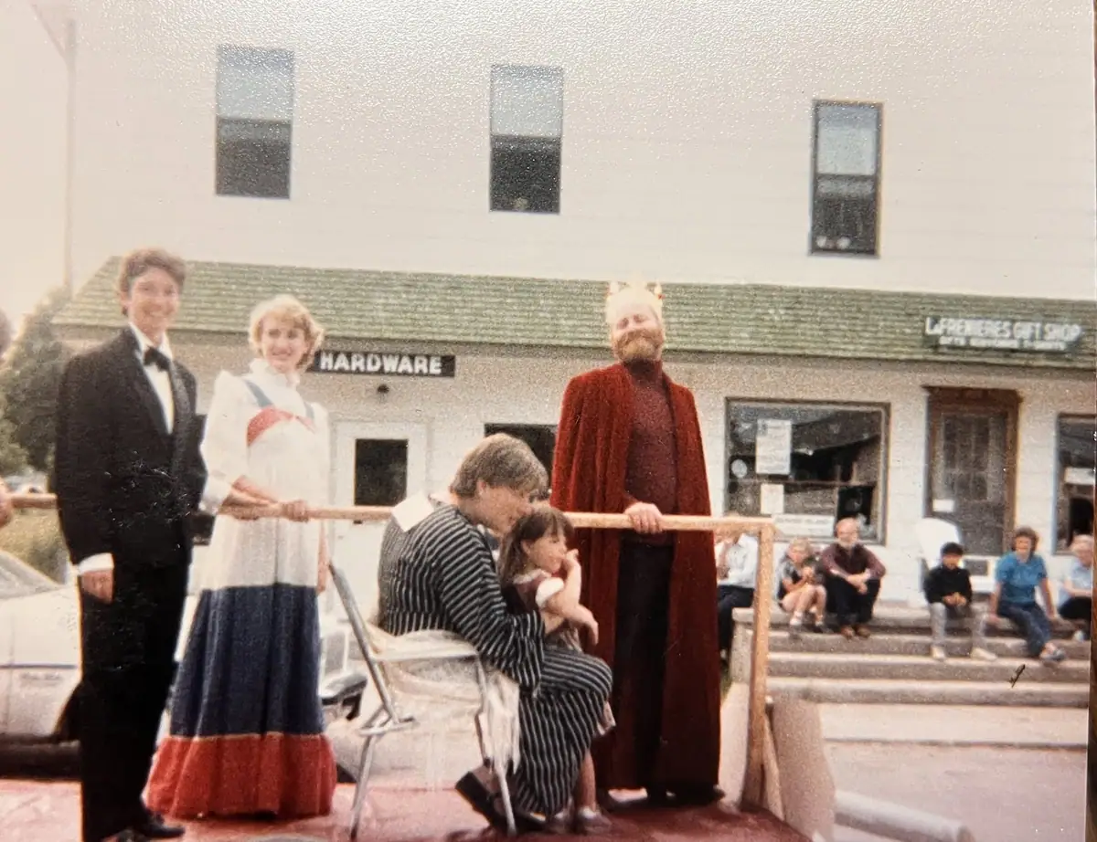 Costumed performers stand on a raised platform before storefronts, with seated spectators nearby.