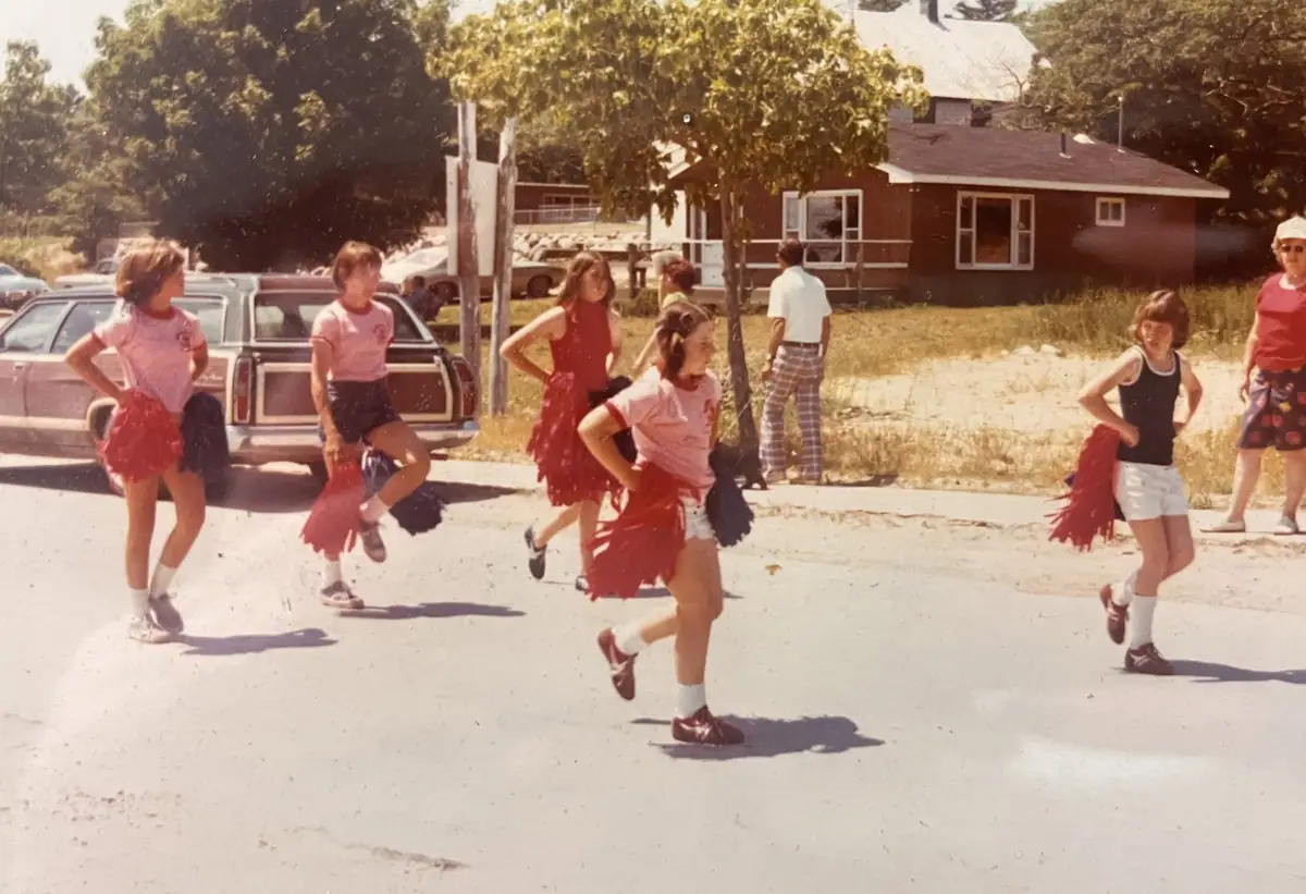 Several young women perform with red pom-poms on a residential street near a house and parked cars.