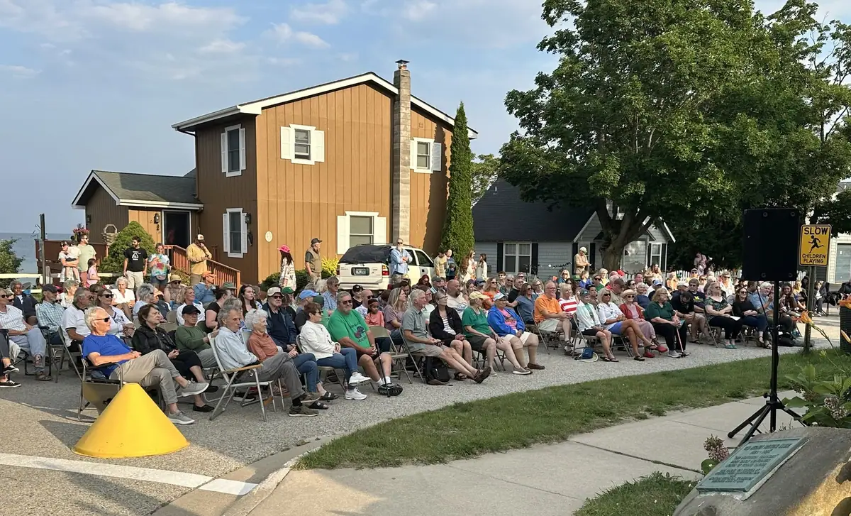 Outdoor gathering with a seated crowd, speaker, house, traffic cone, and roadside warning sign.