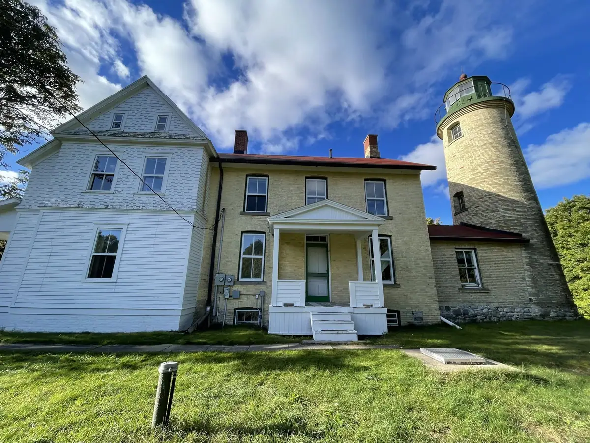 A brick lighthouse and attached white-and-brick keeper’s dwelling stand beside a grassy lawn under a cloudy blue sky.