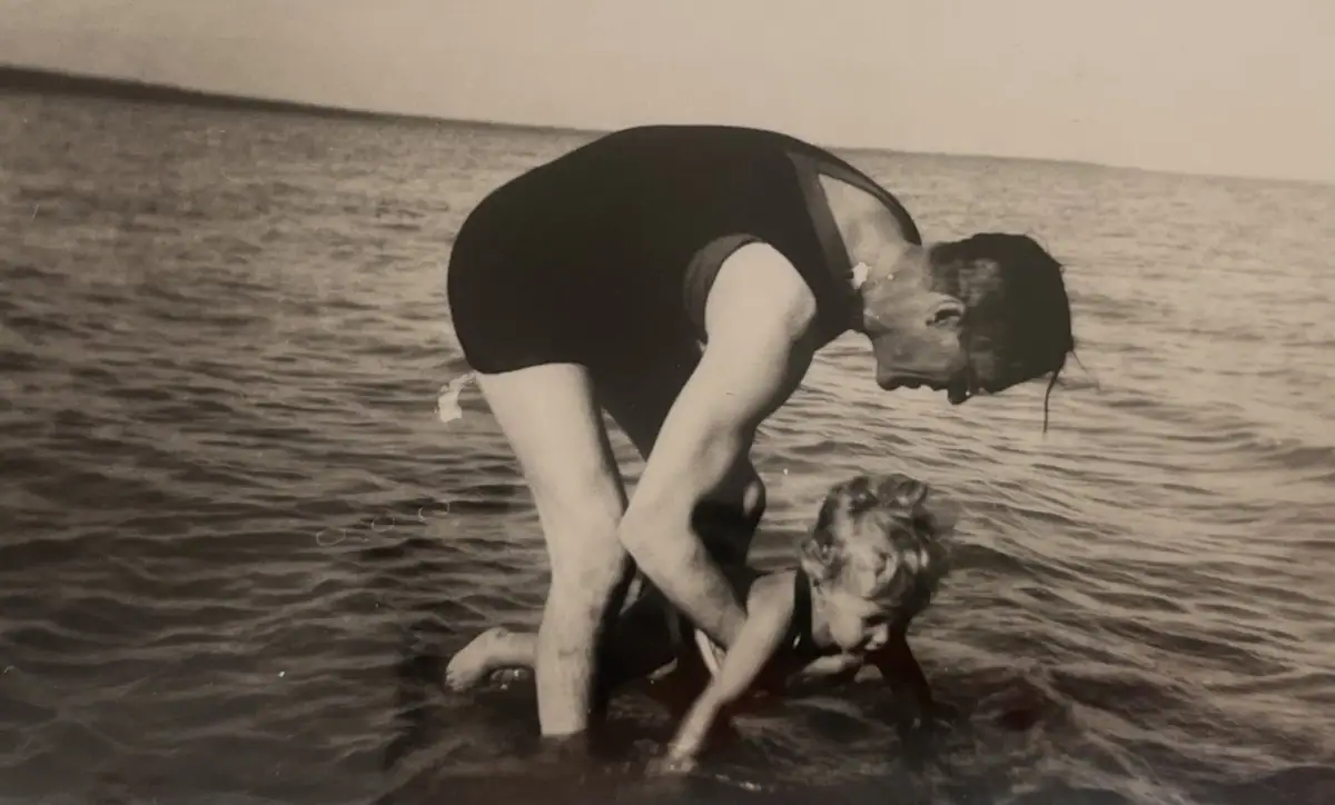 A woman in a dark swimsuit bends over a young child in shallow water.