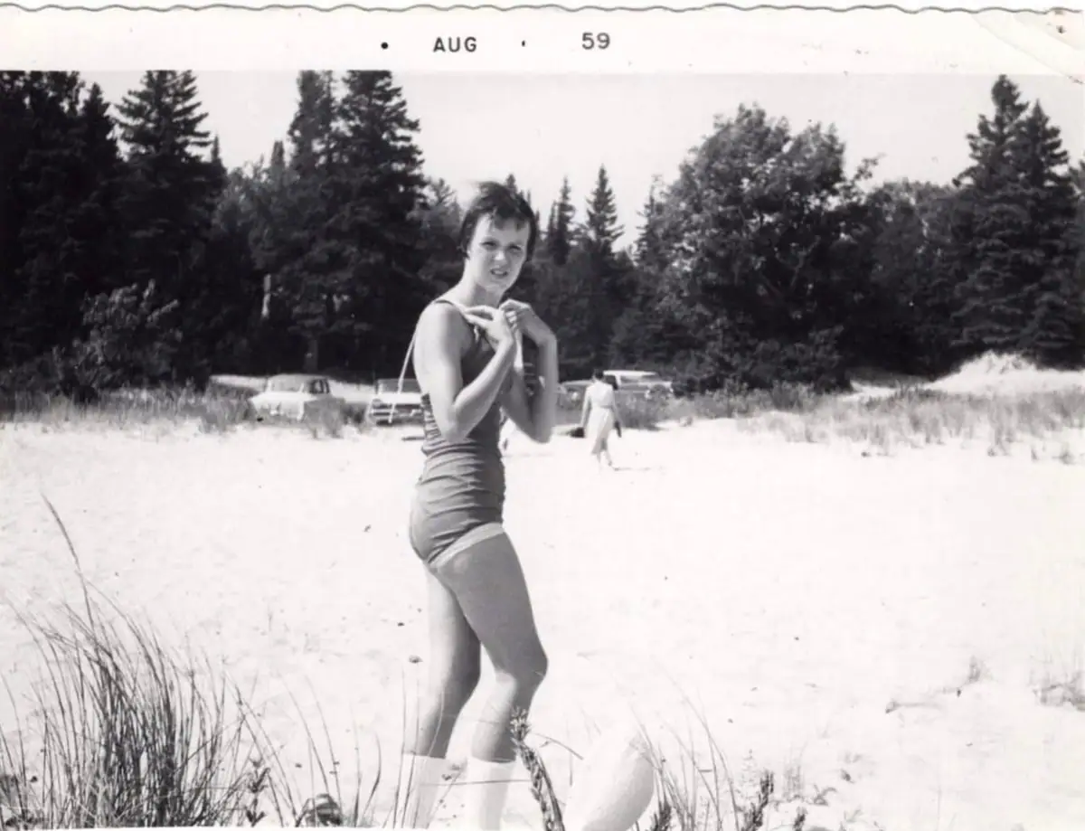 A woman in a one-piece swimsuit poses on a sandy beach beside dunes, trees, parked cars, and another beachgoer.
