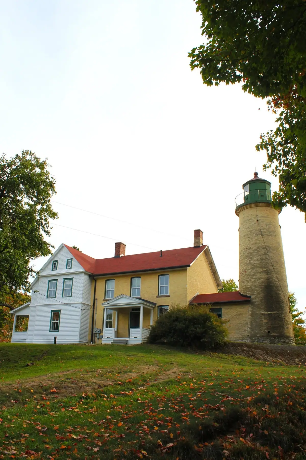 A brick lighthouse and adjoining yellow keeper’s house stand on a grassy slope beneath autumn trees.