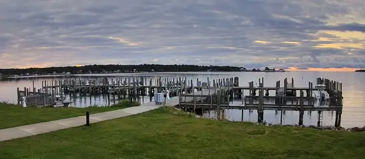 A grassy waterfront with a concrete walkway, wooden docks, and calm water beneath a cloudy sunset sky.