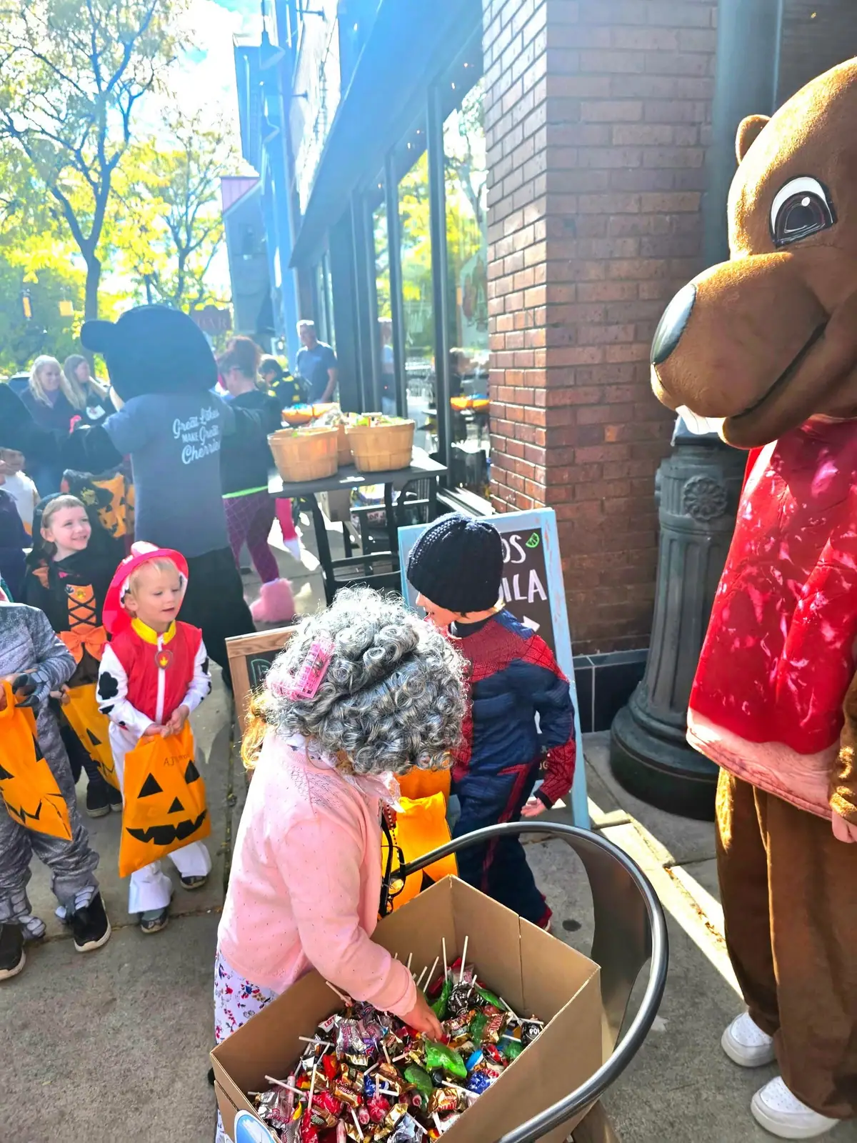 Children in Halloween costumes collect candy beside a large bear mascot outside a storefront.