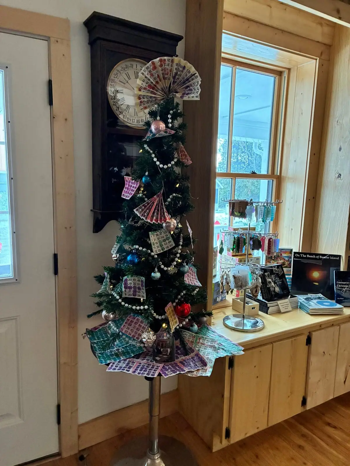 A decorated evergreen tree covered with bingo cards stands beside a tall clock and a wooden merchandise counter.