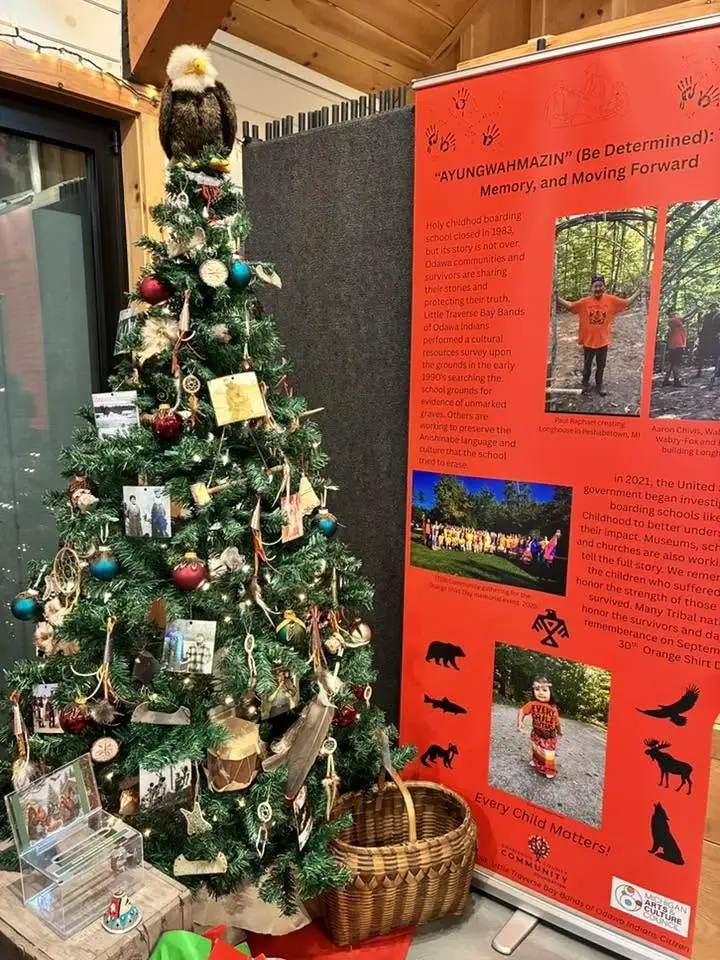 A decorated Christmas tree stands beside a red cultural information display inside a wood-paneled room.