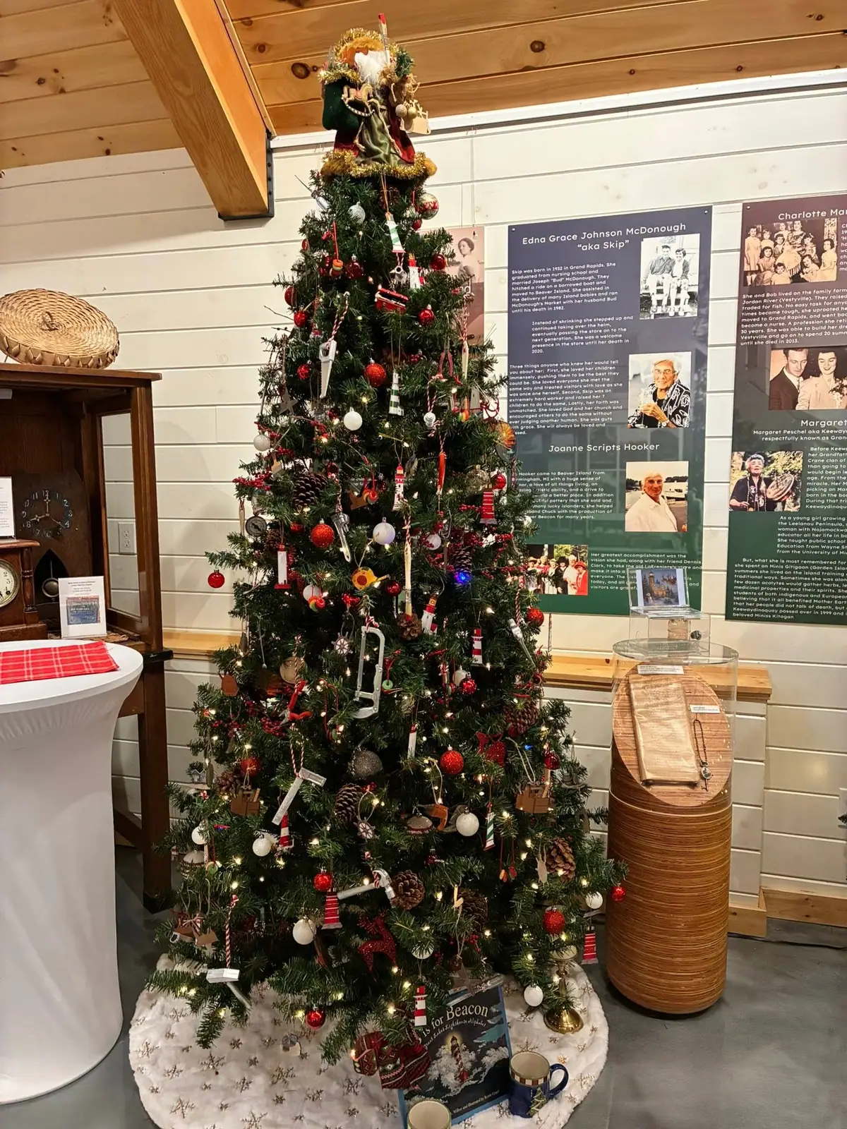 A decorated Christmas tree stands before biographical exhibit panels inside a wood-paneled building.