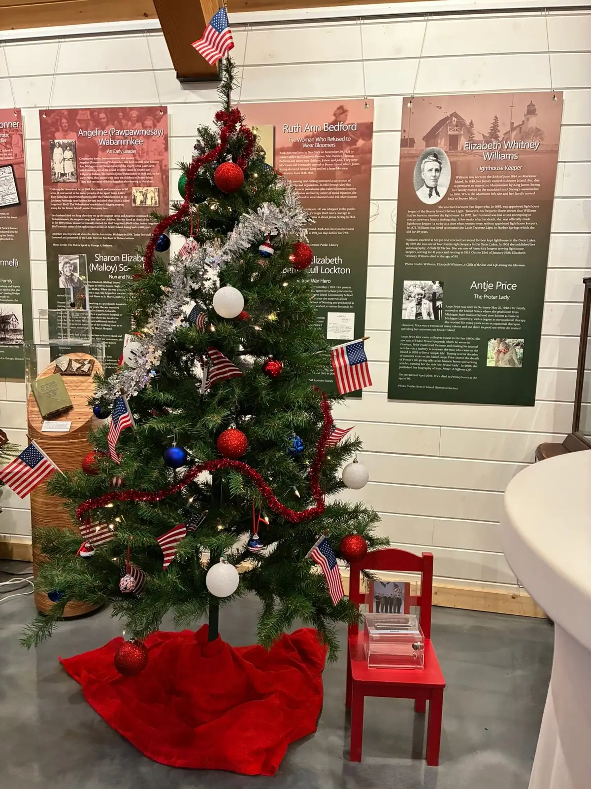 A decorated Christmas tree with American flags stands before historical exhibit panels and a small display chair.