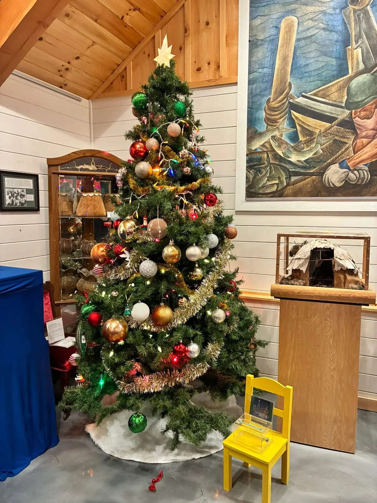 A decorated Christmas tree stands in a wood-paneled room beside a fishing mural and small museum displays.