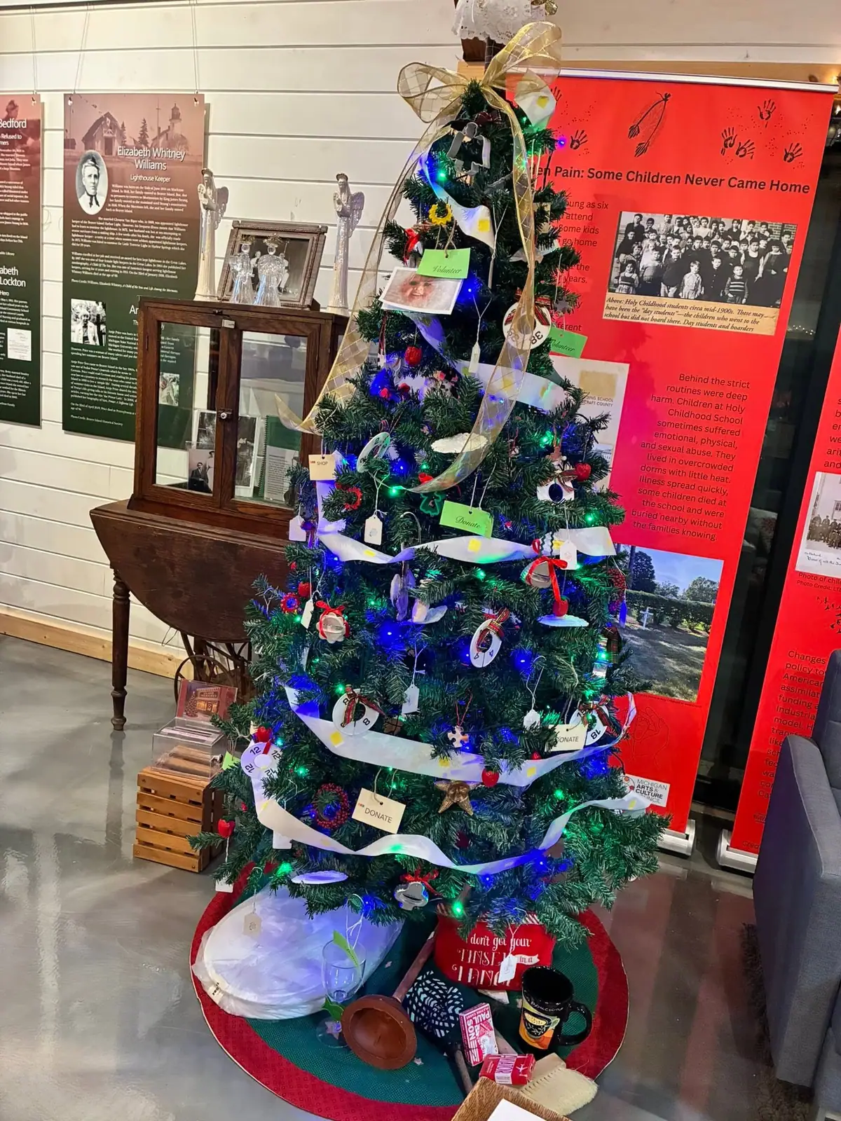 A decorated Christmas tree with lights and donation tags stands before historical exhibit panels and a glass-front cabinet.