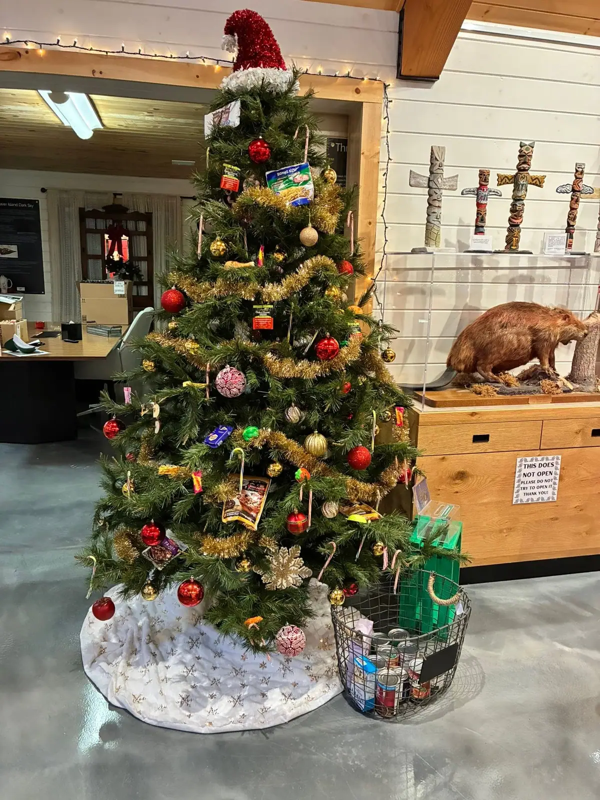 A decorated food-donation Christmas tree stands inside a room beside a wire basket of canned goods.