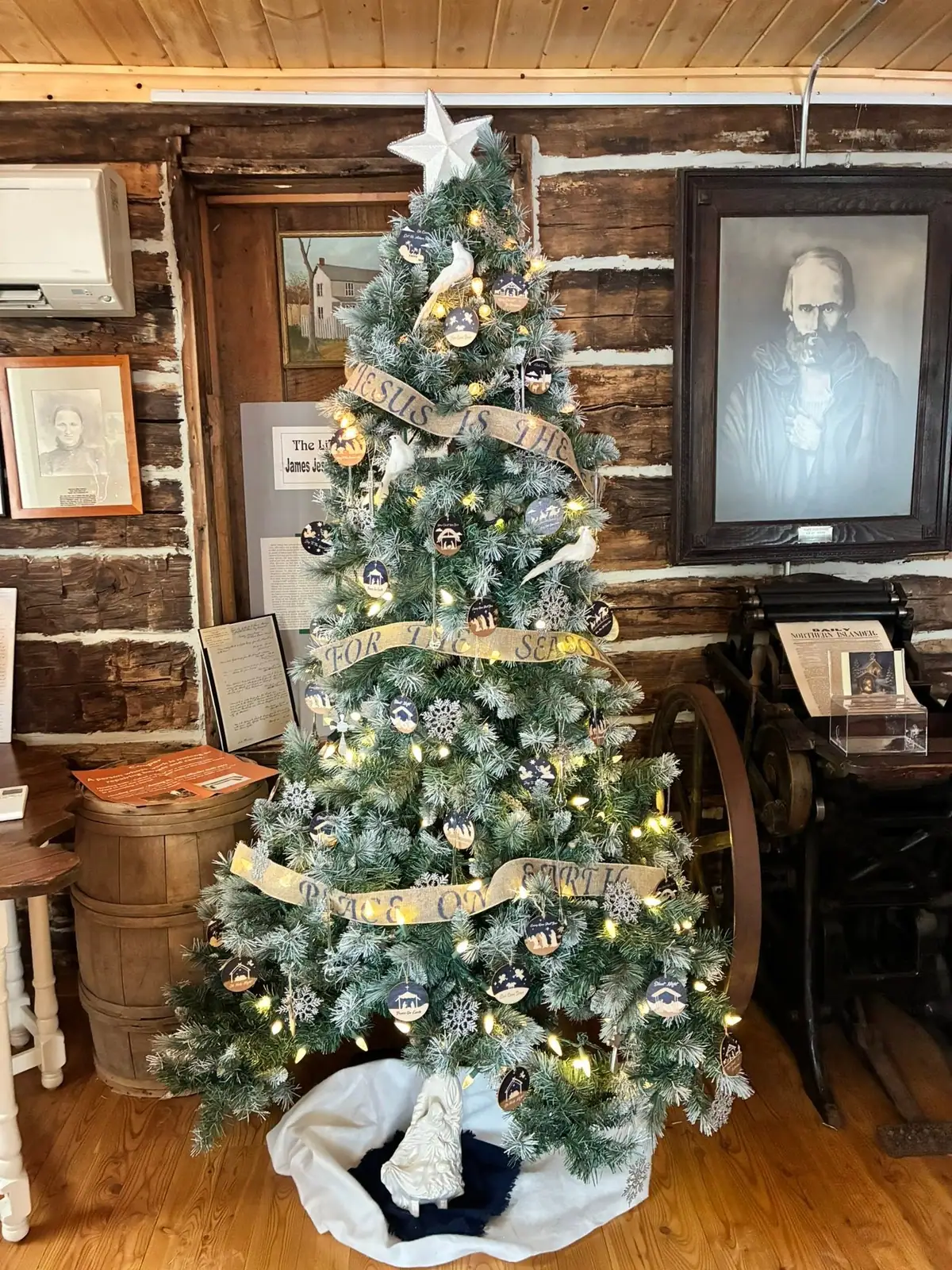 A decorated Christmas tree stands in a rustic log-walled room surrounded by framed portraits and historical displays.