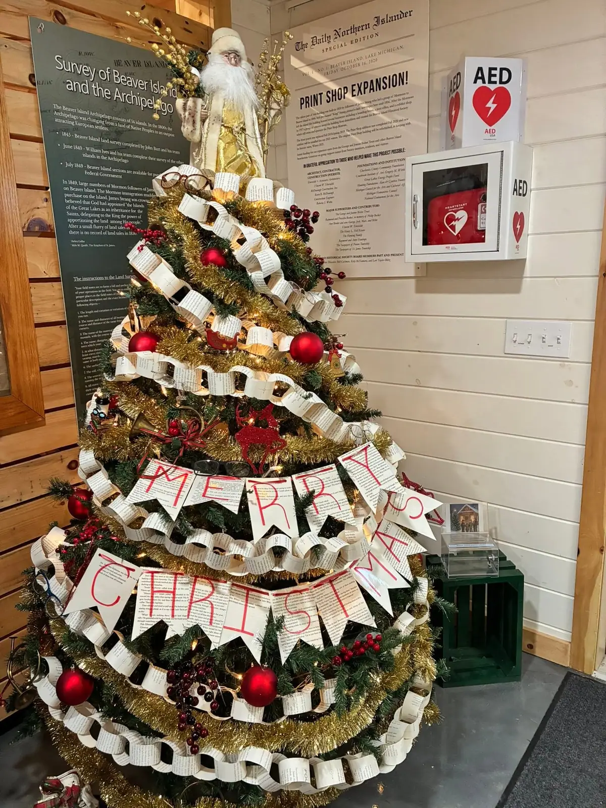 Decorated Christmas tree with a MERRY CHRISTMAS banner beside historical displays and an AED cabinet.