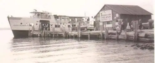 A large ferry is moored beside a wooden pier and dock building, with several people nearby.