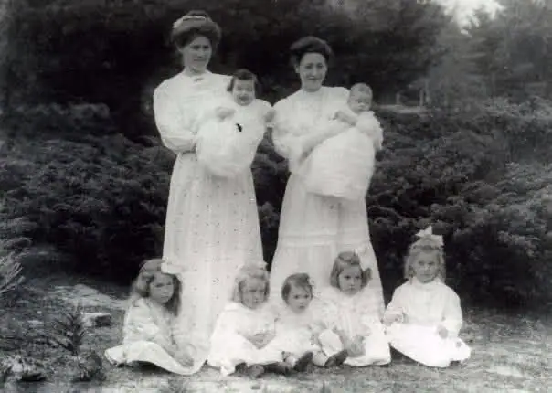 Two women holding babies stand behind five seated children in a posed outdoor portrait.