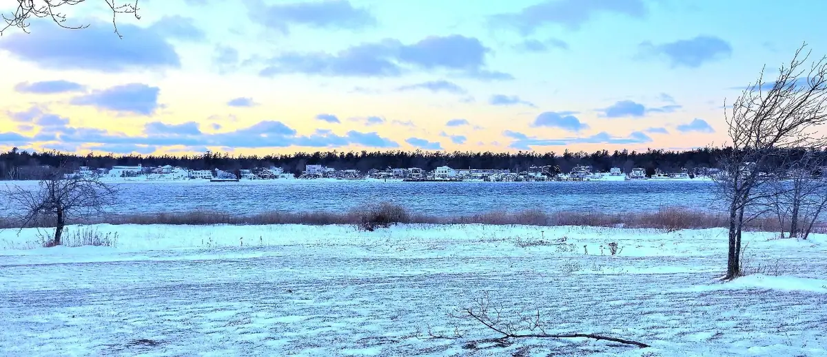 Snow-covered field and shoreline facing distant houses and trees beneath a colorful winter sky.