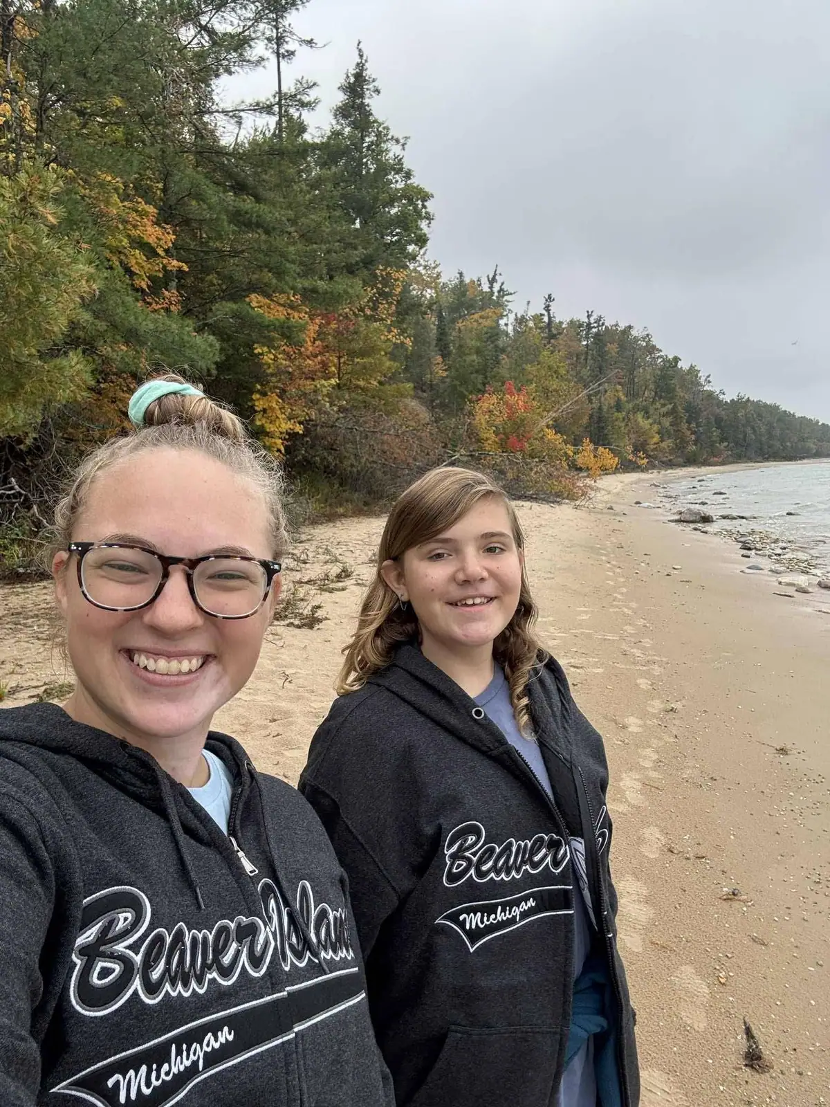 Two people pose on a sandy shoreline with autumn trees and shallow water behind them.