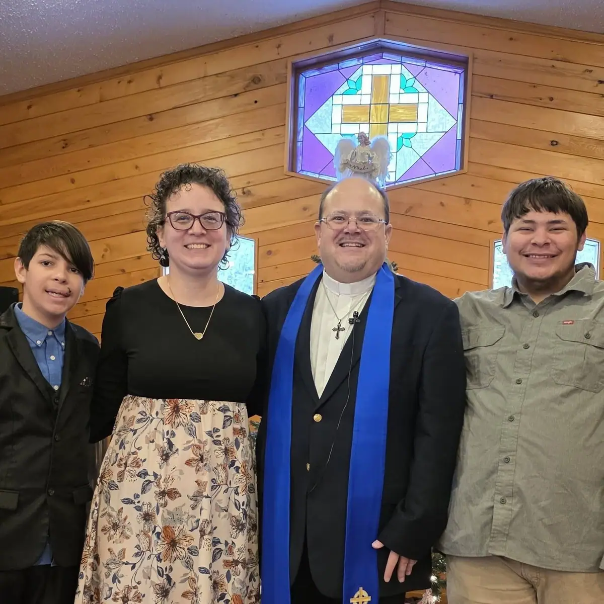 Four people pose together indoors beneath a stained-glass cross.