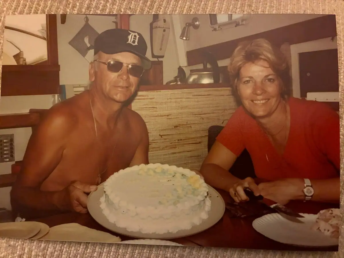 Two adults sit at a table behind a large decorated cake in a warmly lit interior.