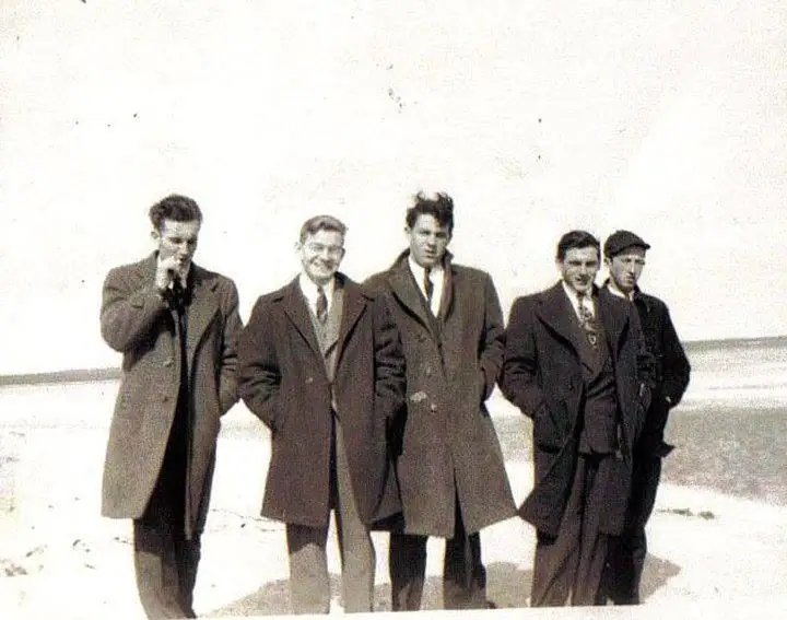 Five men in heavy coats stand together on a sandy shoreline with calm water behind them.