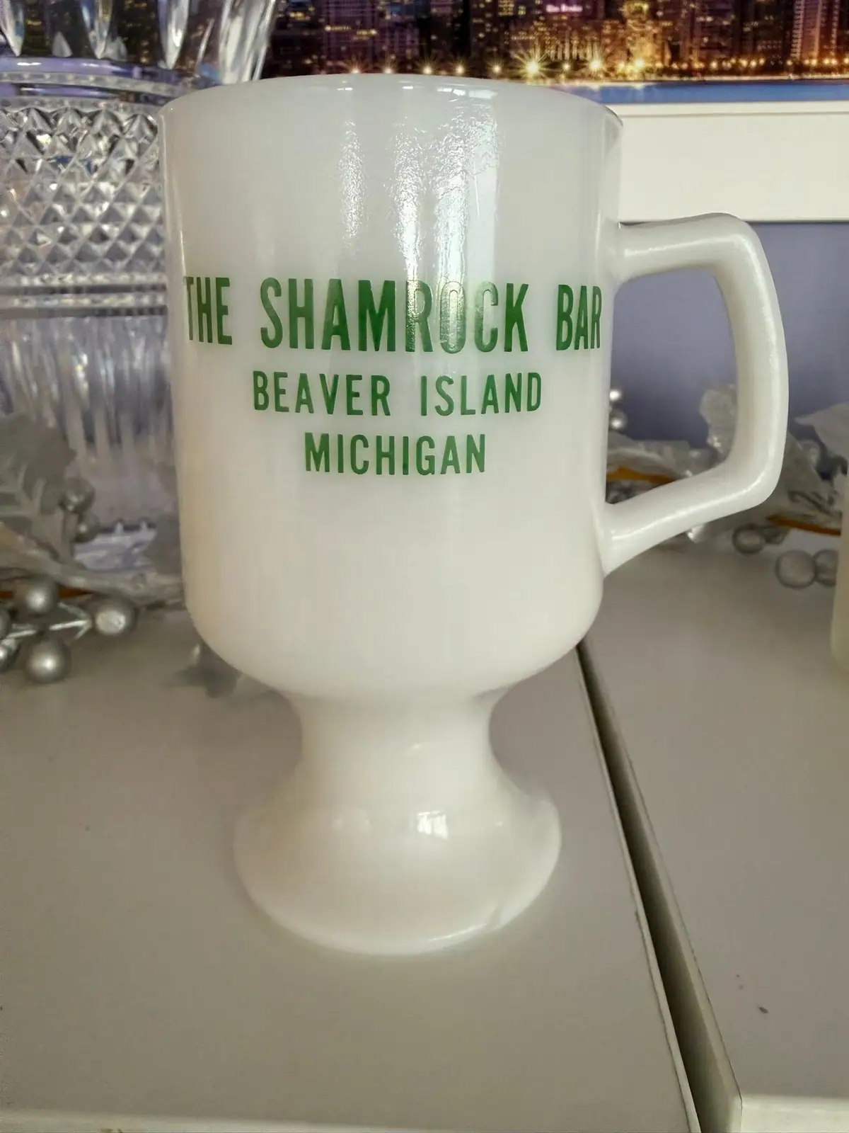 White pedestal mug with green lettering displayed among glassware.