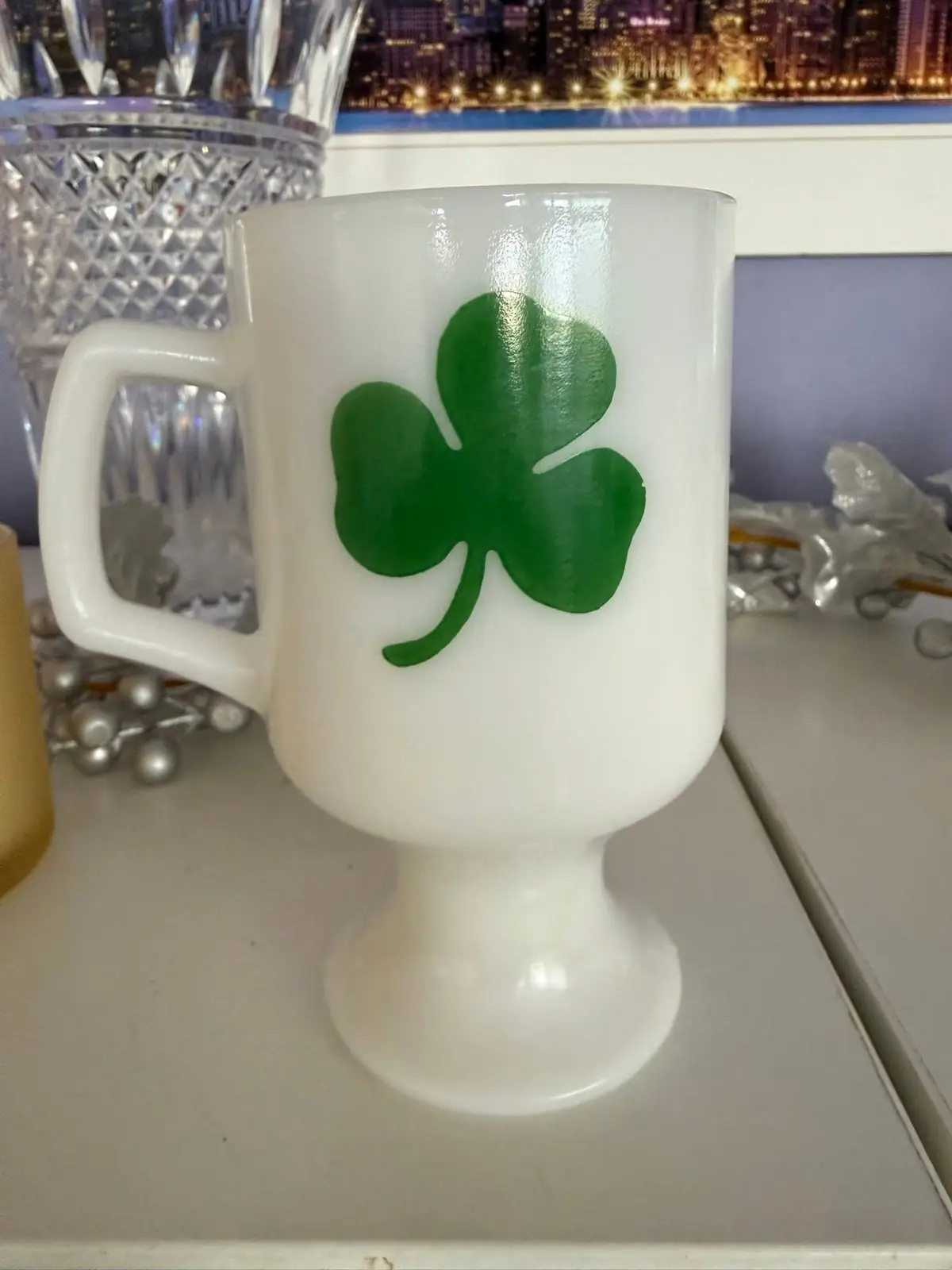 A white pedestal glass mug with a green shamrock sits on a tabletop beside a cut-glass vessel.