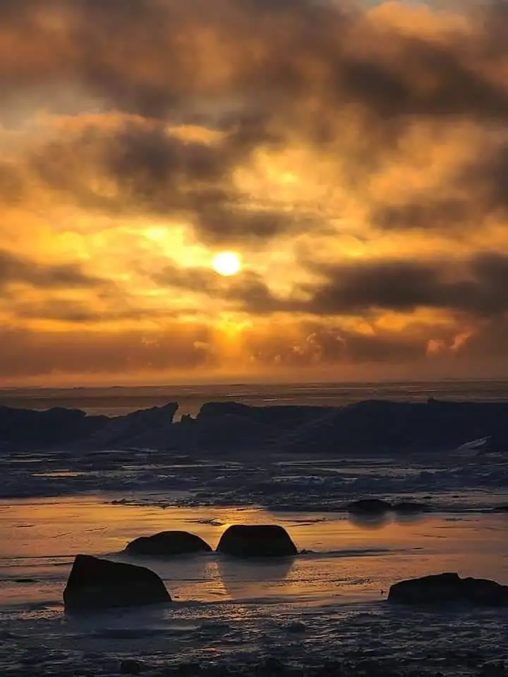 Sunset over a rocky shoreline with dark clouds, reflective water, and silhouetted rocks.