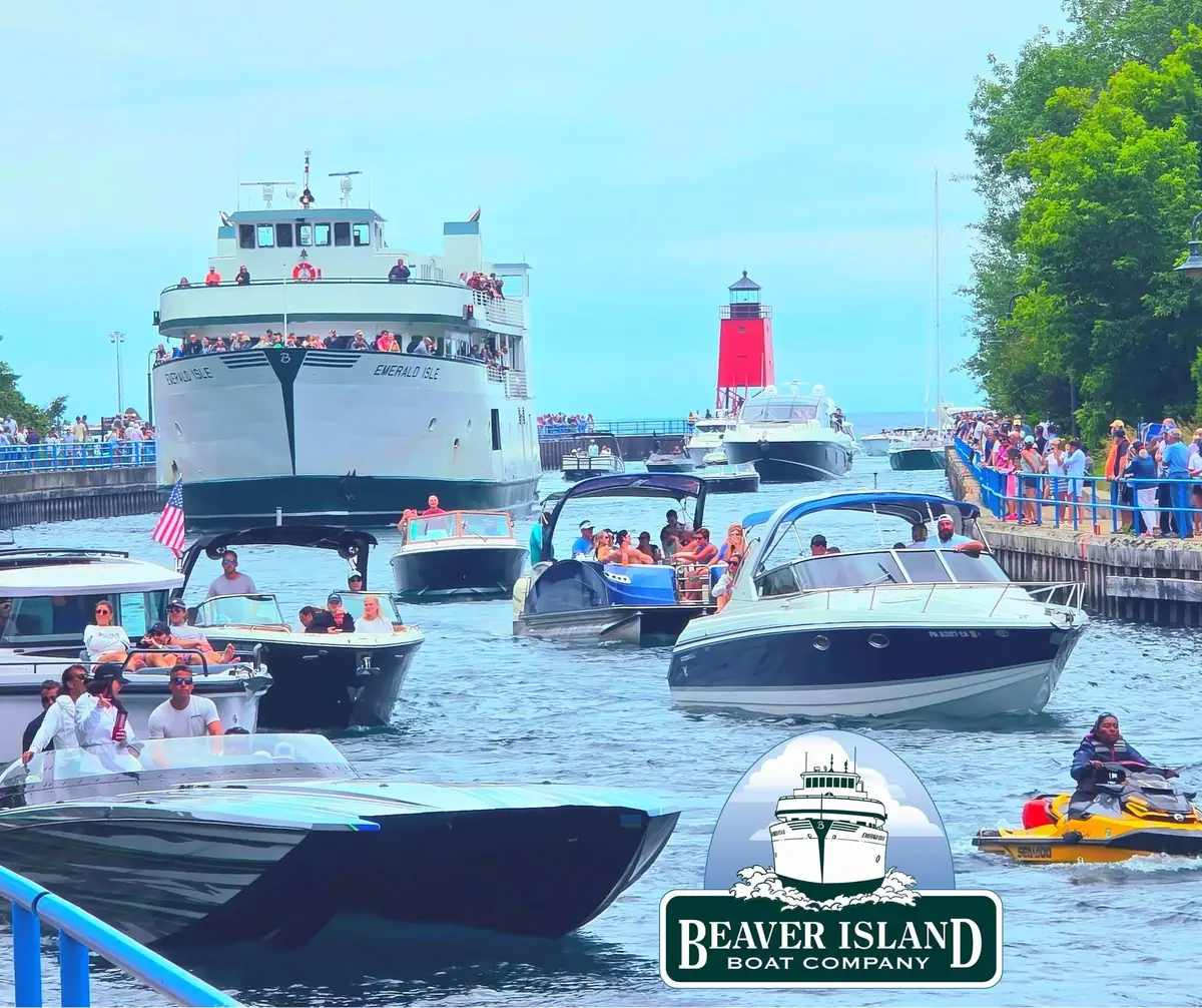 A passenger ferry enters a crowded harbor beside a red lighthouse and numerous recreational boats.