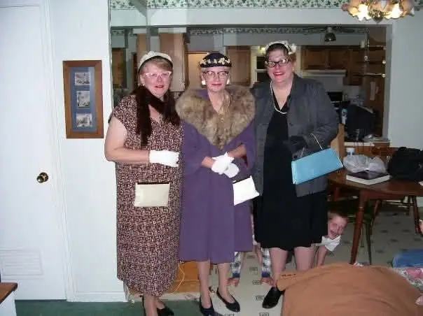 Three women in vintage-style clothing pose indoors, wearing gloves and carrying small handbags.