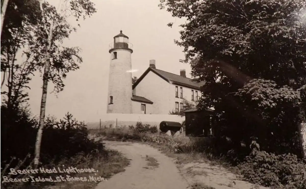 Sepia photograph of a cylindrical lighthouse and attached building beyond a path lined with trees.