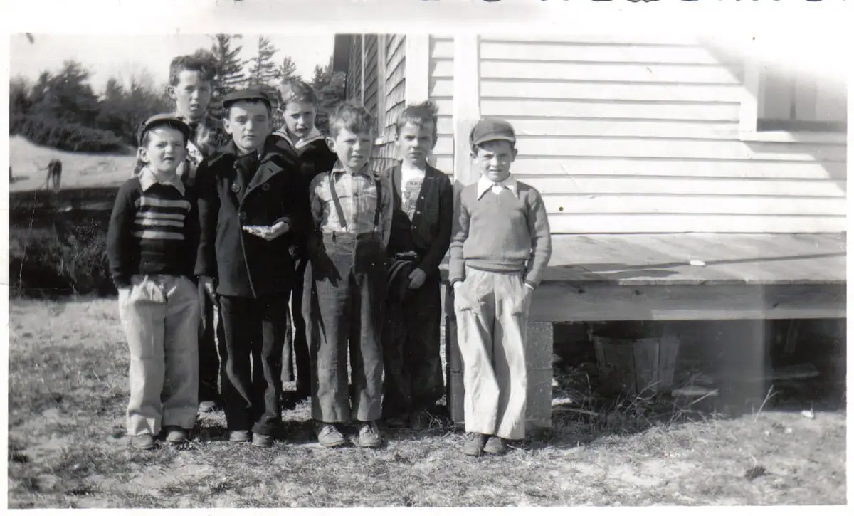 Seven boys pose beside a wooden building in a black-and-white photograph.