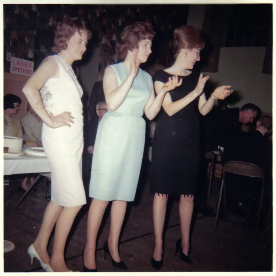 Three women in dresses gesture together at a seated social gathering beside a sign reading “LADIES UPSTAIRS.”