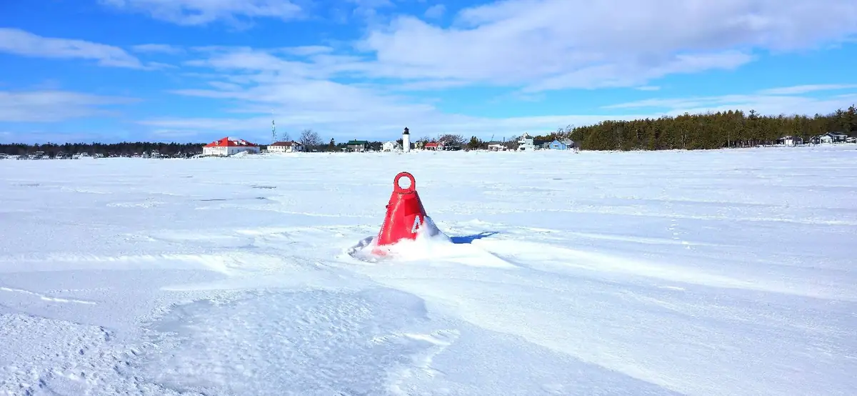 A red navigation buoy protrudes from snow-covered ice, with a lighthouse and shoreline buildings in the distance.