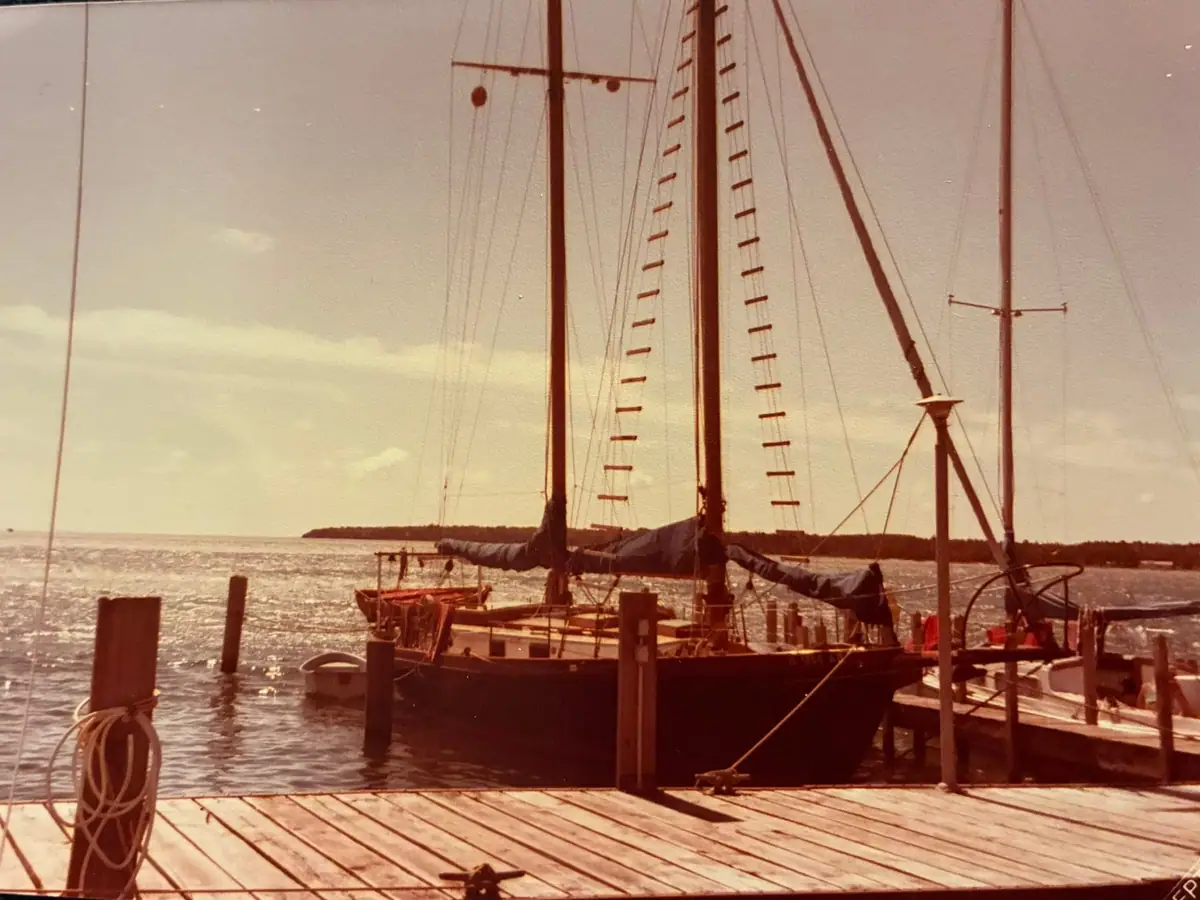 Two-masted sailing vessel moored beside a wooden dock, with rigging, coiled rope, and a distant shoreline.