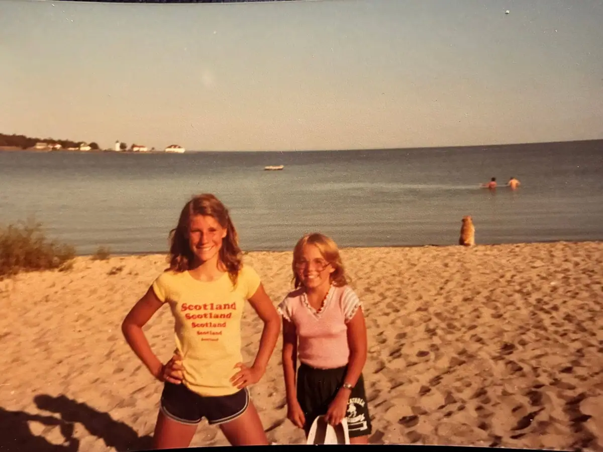 Two women pose on a sandy beach with swimmers and a small boat in the water behind them.