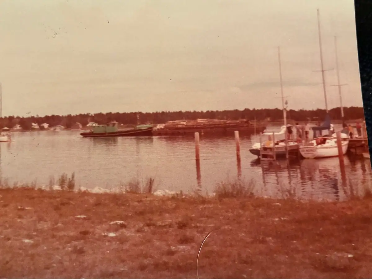 Vintage-tinted harbor photograph showing a tugboat, log-laden barge, and sailboats at a dock.
