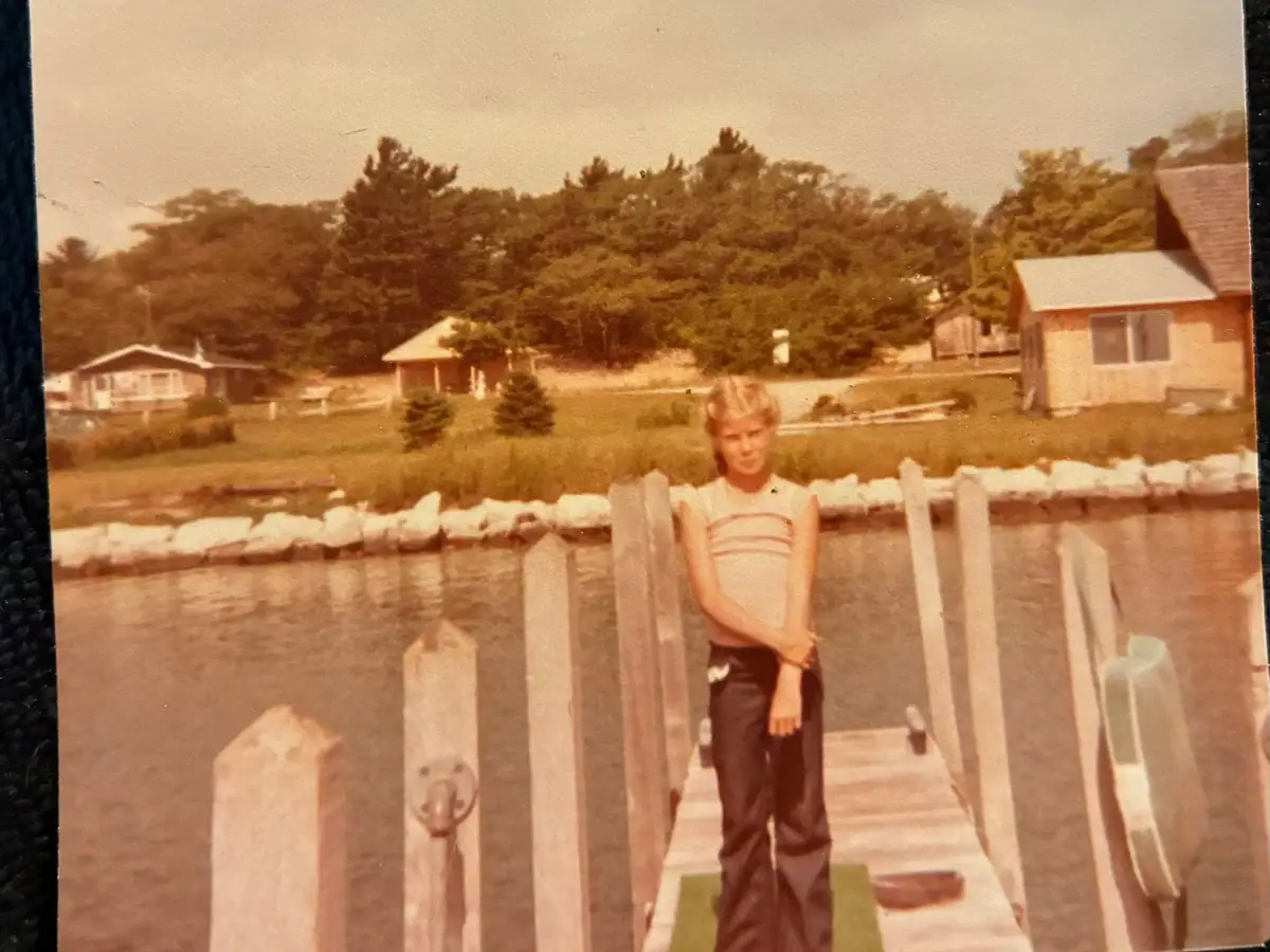 A child stands on a wooden dock beside a white fence, with homes, trees, and rocky shoreline behind.