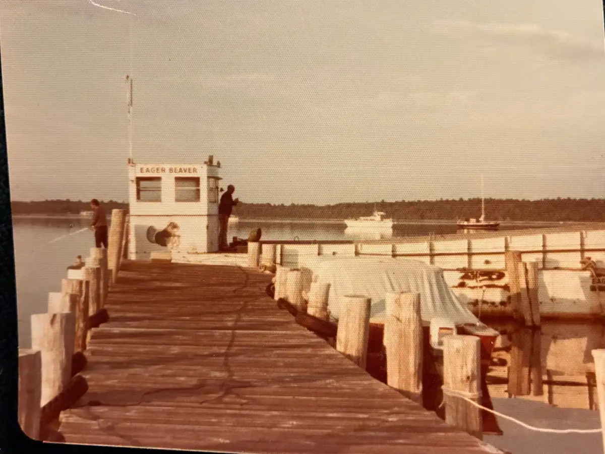 A wooden pier leads to a small white boat named EAGER BEAVER, with two people aboard or nearby.
