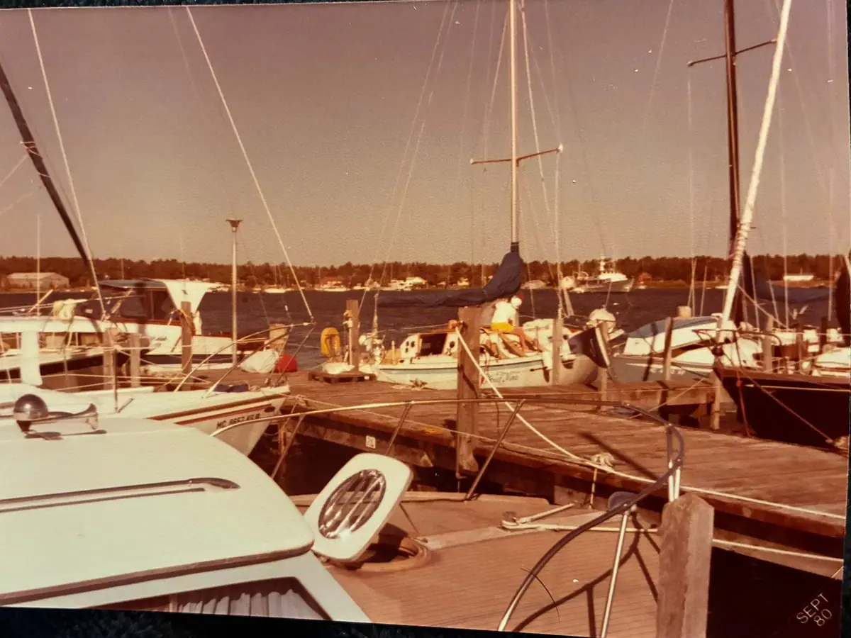 Boats and sailboats moored along a wooden dock, viewed from the deck of another boat.